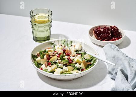 Obstsalat mit Nüssen, ausgewogenes Essen, sauberes Essen. Spinat mit Äpfeln, Pekannüssen und Feta, garniert mit Granatapfelkernen in einer Schüssel auf dem Tisch mit Weißwein Stockfoto