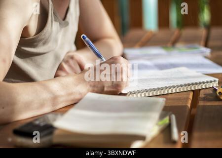 Nahaufnahme der Hand einer Frau mit einem Stift, schreibt in ein Notizbuch, legte ein Buch auf den Tisch, Handy, Bleistifte, draußen auf einer Terrasse Stockfoto