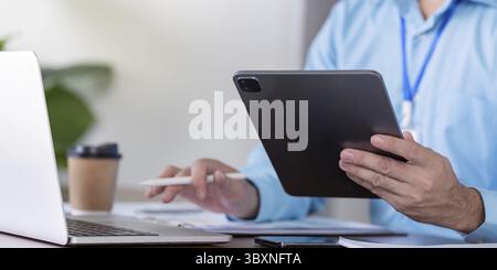 Ein Geschäftsmann, der ein Tablet und einen Stift verwendet, während er an einem Laptop arbeitet und sich auf die digitale Planung in einer modernen Büroumgebung konzentriert Stockfoto
