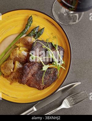 Appetitliche Mahlzeit aus gegrilltem Schweinefleisch mit zartem geröstetem Spargel, gebackener Kartoffel in Haut und cremiger Sauce, serviert auf sonnigem gelben Teller mit einem Glas Stockfoto
