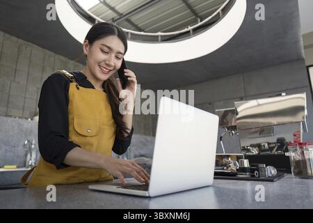 Lächelnde junge Barista in gelber Schürze, Multitasking mit Laptop und Telefon in einem modernen Café Stockfoto