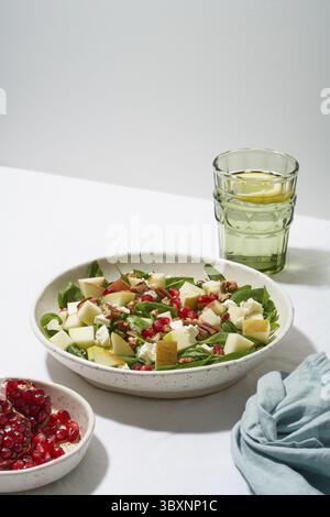 Obstsalat mit Nüssen, ausgewogenes Essen, sauberes Essen. Spinat mit Äpfeln, Pekannüssen und Feta, garniert mit Granatapfelkernen in einer Schüssel auf dem Tisch mit Weißwein Stockfoto