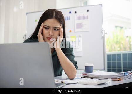 Eine überforderte, gestresste Geschäftsfrau, die mit einem Laptop arbeitet und gleichzeitig Kopfschmerzen im Büro hat Stockfoto