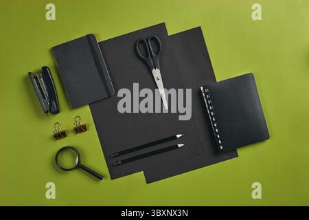 Zwei Blatt schwarzes Papier, zwei Bleistifte, Ordner, Hefter, Notebook, Schere, goldenen Büroklammern und Lupe auf grünem Hintergrund mit Kopie Raum. Edu Stockfoto
