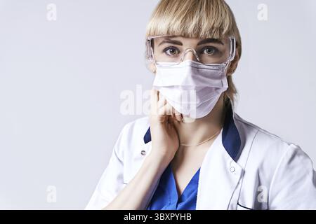 Selbstbewusste blonde Ärztin mit Schutzmaske. Porträt auf hellgrauem Hintergrund Stockfoto