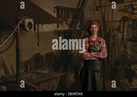 Professioneller Schmied im Schmiedeporträt. Junge lächelnde europäische Frau trägt Uniform und überkreuzte Arme. Idee der handgefertigten Metallarbeit. Das Konzept des Handwerks Stockfoto
