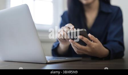Eine junge Geschäftsfrau am Telefon im Büro. Geschäftsfrau, die am Telefon SMS schreibt und am Laptop arbeitet. Hübsche junge Geschäftsfrau, die auf der Arbeitsplatte sitzt Stockfoto