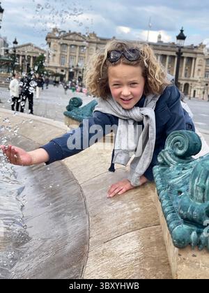 Das lockhaarige Kind streckt sich aus, um das fließende Wasser eines dekorativen Brunnens auf einem geschäftigen Platz zu berühren und zeigt Neugier und Freude. Gebäude und Menschen Stockfoto