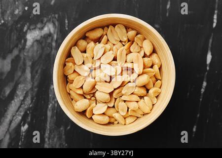 Blick von oben auf gesalzene Erdnüsse in einer Schüssel. Köstlicher Snack für Party oder Bar. Leckere geröstete Nüsse auf schwarzem Marmortisch. Knusprige Textur und goldene Farbe. Stockfoto