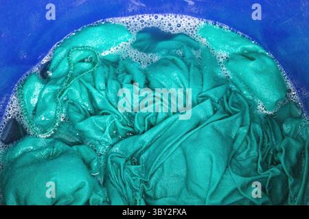 Nahaufnahme eines türkisfarbenen Stoffes, das in einem Kunststoffeimer in sprudelndem Waschmittelwasser getaucht ist und leuchtende Farben und Schaumstoffe zeigt. Ideal zum Waschen, Reinigen Stockfoto
