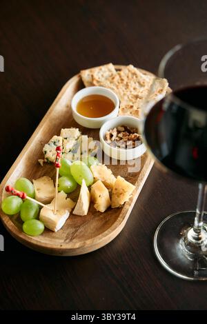 Eine luxuriöse Holzplatte bietet verschiedene Käsesorten, saftige Trauben, Honig und Nüsse, perfekt kombiniert mit einem Glas reichen Rotwein für einen exquisiten Tag Stockfoto