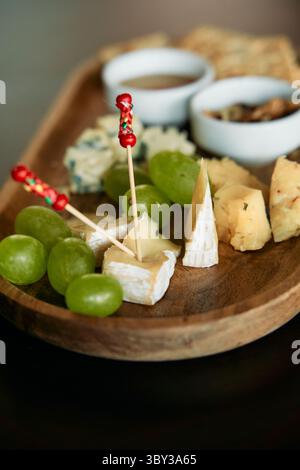 Eine elegante Platte mit verschiedenen Gourmet-Käsesorten, frischen grünen Trauben, verschiedenen Nüssen und Dips, kunstvoll auf einer hölzernen Servierplatte angeordnet Stockfoto
