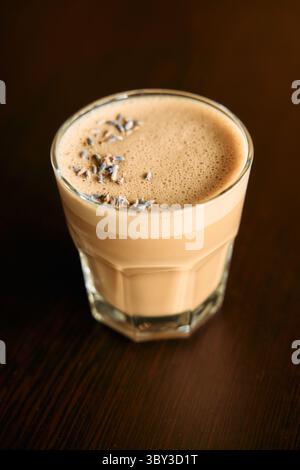 Ein cremiger Lavendellatte in einem klaren Glas sitzt auf einem Holztisch und bietet eine schaumige Platte mit Lavendelstreuseln für ein wohltuendes aromatisches Erlebnis Stockfoto