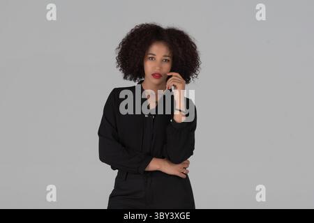 Porträt einer stilvollen jungen afroamerikanischen Frau mit lockigen Haaren und schwarzem Outfit, die selbstbewusst das Kamerastudio auf grauem Hintergrund betrachtet. Stockfoto