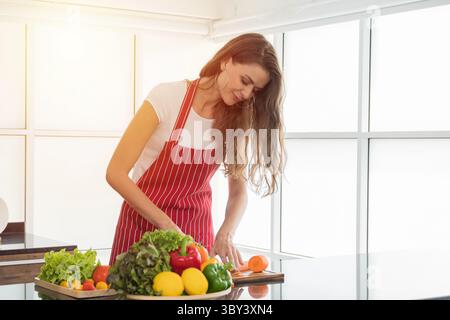 Frau in einer roten Schürze zerkleinert präzise frische Karotten auf einem Holzbrett, mit einer lebendigen Schüssel mit Gemüse in der Nähe, was die gesunde Ernährung in der Küche betont. Stockfoto