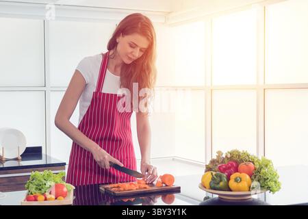 Frau in einer roten Schürze zerkleinert präzise frische Karotten auf einem Holzbrett, mit einer lebendigen Schüssel mit Gemüse in der Nähe, was die gesunde Ernährung in der Küche betont. Stockfoto