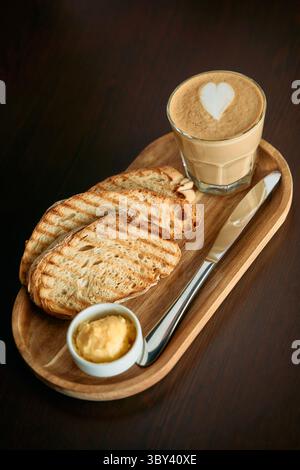 Eine gemütliche Frühstücksszene mit einem Cappuccino mit herzförmiger Latte Art, serviert neben gegrilltem Brot und Butter auf einem Holztablett mit einem Messer, pe Stockfoto
