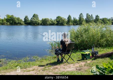 Junger Mann mit langen Haaren fischen an einem sonnigen Tag am Fluss mit grünem Gras und Wasserpflanzen Stockfoto
