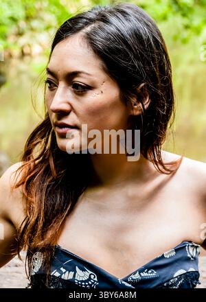 Eine Frau mit dunklen Haaren und einem Tattoo am Hals schaut zur Seite. Sie trägt ein dunkelblaues Kleid mit Schmetterlingsmuster. Stockfoto