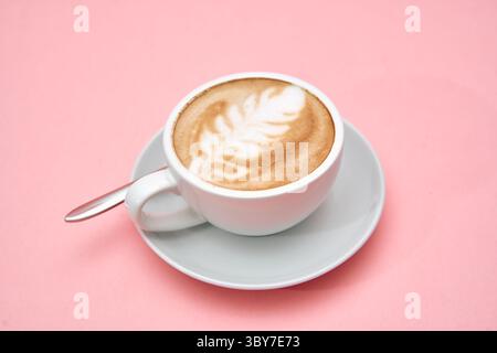 Ein wunderschön gefertigter Latte mit Blattkunst sitzt in einer weißen Tasse auf einer sanften rosa Oberfläche und zeigt das komplizierte Design und die einladende Eleganz des Fro Stockfoto