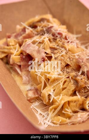 Cremige Carbonara garniert mit Parmesan und schwarzem Pfeffer, serviert in einer praktischen Take-away-Box auf rosafarbener Oberfläche, die die Köstlichkeit italiens unterstreicht Stockfoto
