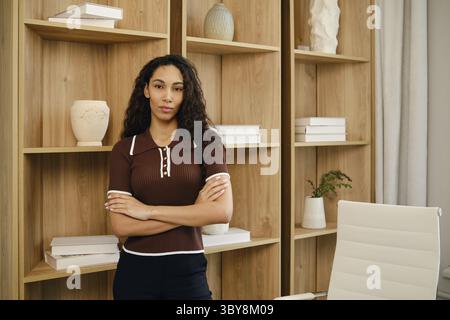 Eine selbstbewusste Geschäftsfrau steht mit überkreuzten Armen in einem modernen Bürobereich. Der Hintergrund ist mit Holzregalen gefüllt mit dekorativen Gegenständen Stockfoto