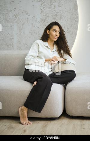 Niedliche Frau mit langen lockigen Haaren sitzt bequem auf einer modernen Couch in einem stilvollen Wohnzimmer Stockfoto