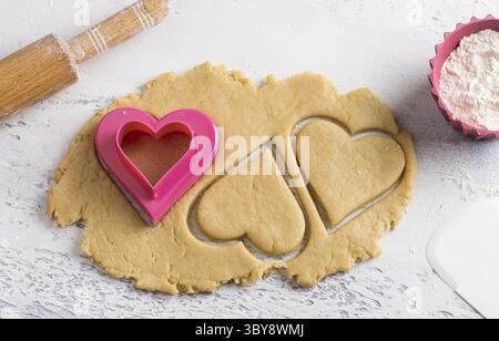Essen, Cutted Heart Shape Teig Plätzchen mit Keksschneider, Nudelnadel und Schüssel Mehl auf einer hellblauen Tischplatte, Blick von oben. Stufe des Kochens von Delikatessen Stockfoto