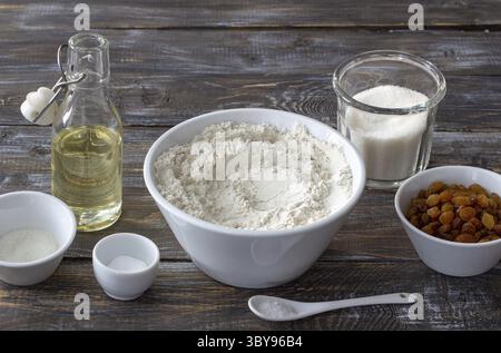 Lebensmittel, Backzutaten einschließlich Mehl, goldene Rosinen, Zucker, Öl, Soda, Vanillezucker und Salz in Schüsseln auf rustikalem Holztisch, für Rezeptkonzept Stockfoto