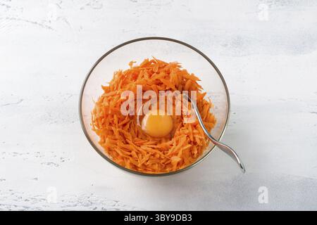Essen, Glasschale mit geriebenen Karotten mit einem Ei auf einem hellgrauen Tisch, Blick von oben. Hausgemachte vegetarische Gerichte: Karottenpfannkuchen, Krapfen oder anderes Stockfoto