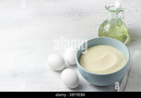 Lebensmittel, Backzutaten: Hellblaue Schüssel mit Grieß, frische weiße Eier, eine Glasschale mit Kristallzucker und eine Flasche Pflanzenöl auf einem weißen Stockfoto