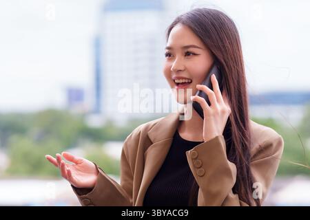 Moderne asiatische Geschäftsfrau führt im Freien ein lebhaftes Smartphone-Gespräch. Ihre ausdrucksstarke Geste vermittelt dynamische Kommunikation und urbane Verbindung Stockfoto