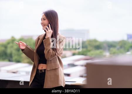 Eine junge asiatische Frau führt ein wichtiges geschäftliches Telefongespräch von einem modernen Stadtbalkon aus, das Professionalität und Selbstvertrauen ausstrahlt Stockfoto
