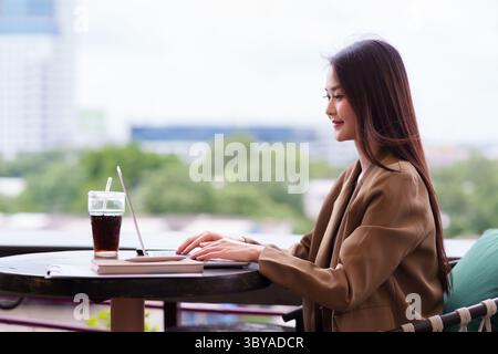 Selbstbewusste asiatische Frau, die im Freien an einem Laptop arbeitet Sie konzentriert sich auf ihre Geschäftsaufgaben und genießt einen flexiblen, modernen Arbeitsstil Stockfoto