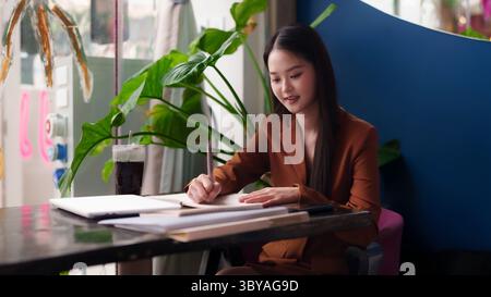 Asiatische Erwachsene Frauen konzentrieren sich darauf, Notizen am Café-Tisch zu schreiben. Professionelles weibliches Planungsprojekt im modernen, inspirierenden Arbeitsbereich. Stockfoto
