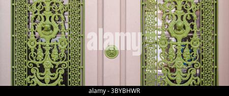 Verzierte grüne schmiedeeiserne Türen mit kunstvollen Mustern und runden Griffen, die sich an einer hellen Wand befinden. Stockfoto
