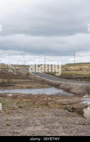 Teriberka, Russland - 13. Juni 2025: Eine abgelegene Straße zieht sich durch die arktische Tundra-Landschaft mit spärlichem Verkehr und Versorgungsmasten entlang des Weges Stockfoto