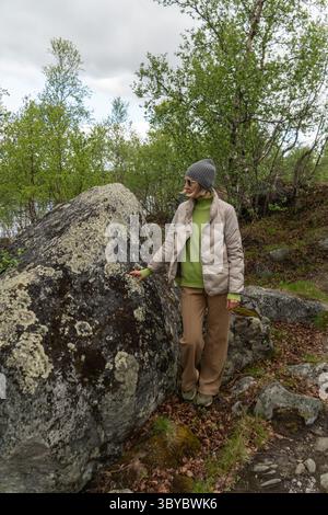 Eine Frau steht in einer Waldlichtung und berührt sanft einen großen, flechtenbedeckten Felsen, während sie neutrale Outdoor-Kleidung und Sonnenbrille trägt Stockfoto