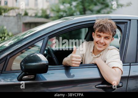 Ein lächelnder junger Fahrer zeigt Daumen hoch und lächelt in die Kamera im Auto. Stockfoto