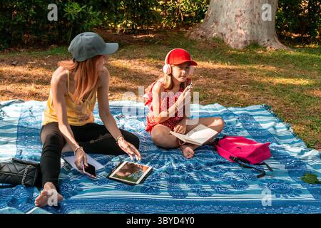 Mutter und Tochter genießen ein entspannendes Picknick und benutzen elektronische Geräte im Park Stockfoto
