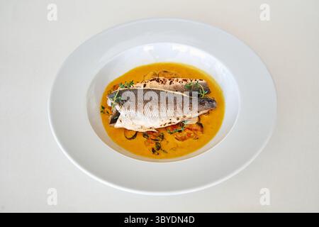 Gegrilltes Wolfsbarschfilet liegt auf einem Bett aus Muscheln in einer reichhaltigen, cremigen Sauce, elegant auf einem weißen Teller serviert, garniert mit Thymian für ein Gourmet-Dinner Stockfoto