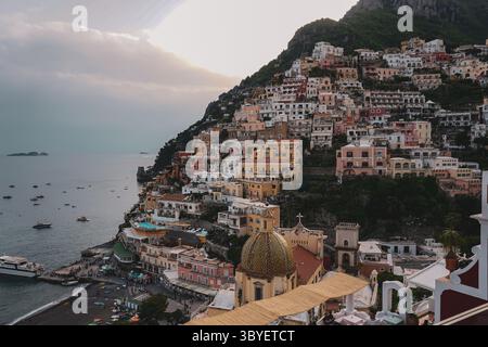 Farbenfrohe Häuser an den Klippen stürzen sich in Positano, Italien, hinunter zum Meer, mit Booten im Hintergrund und üppig grünen Bergen, die dahinter aufsteigen Stockfoto