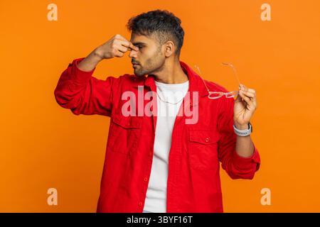Erschöpfter, müder indischer junger Mann zieht die Brille aus, spürt Augenschmerzen, ist überlastet Burnout von langer Arbeit. Verschlafener, erschöpfter Arabianer, der die Augen isoliert auf einem orangen Hintergrund des Studios reibt Stockfoto
