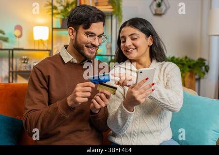 Glückliches hispanisches Paar beim Online-Shopping, indem es Kreditkartennummer auf dem Smartphone eingibt, während es zu Hause auf dem Sofa sitzt. Lächelnde Familienbefriedigung Stockfoto
