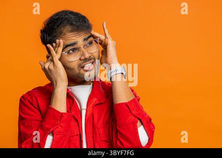 Unzufrieden erschöpfter indischer Hindu-Mann, der Schläfen reibt, um Kopfschmerzen zu heilen, leidet unter Spannung, Migräne, Stress, grimmigen Schmerzen, Bluthochdruck isoliert auf orangefarbenem Hintergrund Stockfoto