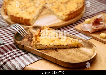 Ein herzhaftes Quiche-Stück mit Schinken und Käse liegt auf einem Holzteller, begleitet von einer Gabel und karierter Serviette, die eine köstliche, appetitliche Atmosphäre schaffen Stockfoto