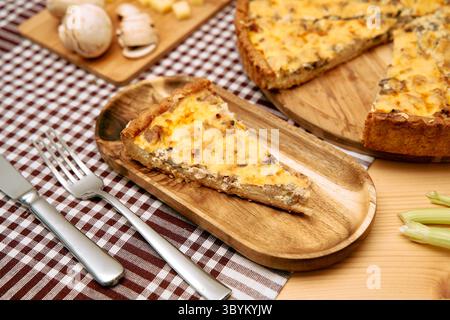 Auf einem Holzteller liegt auf einer karierten Tischdecke ein Stück Pilz- und Käsequiche, die Pilze und Zutaten sichtbar sind und zum Genießen bereit sind. Stockfoto