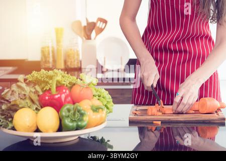 Eine Close-up-Frau in einer roten Schürze zerkleinert präzise frische Karotten auf einem Holzbrett, mit einer lebendigen Schüssel mit Gemüse in der Nähe, die eine gesunde Ernährung betont. Stockfoto