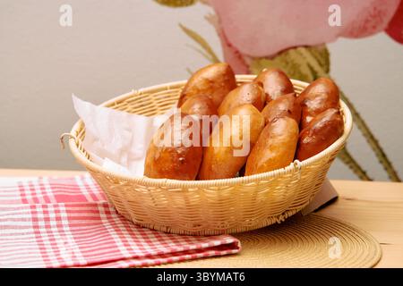 Ein Korbkorb mit frisch gebackenen, goldbraunen Brötchen steht auf einem dekorativen Tisch mit rot-weißem Tuch und floralem Hintergrund Stockfoto