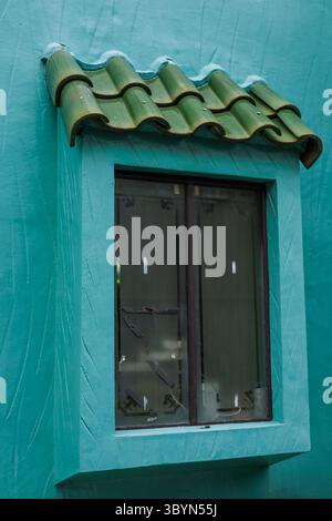 Linker Winkel der Stuckwand in türkisfarbener Farbe, verziert mit einem hellgrünen Kachelüberhang und dunklen Fensterrahmen. Stockfoto